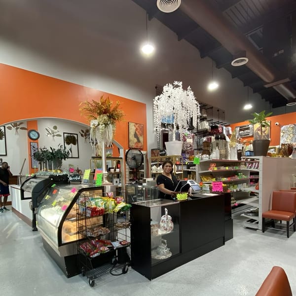 A woman stands behind a counter in a brightly lit store with baked goods and shelves of items.